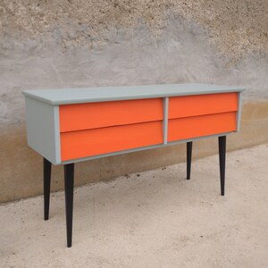 Das Bild zeigt eine original Vintage Kommode mit vier orange gestrichenen Schubladen. Der Korpus ist grün, und die langen Holzbeine sind schwarz gestrichen. Die Kommode wird leicht seitlich von links präsentiert. Blickfang mit Retro-Charme