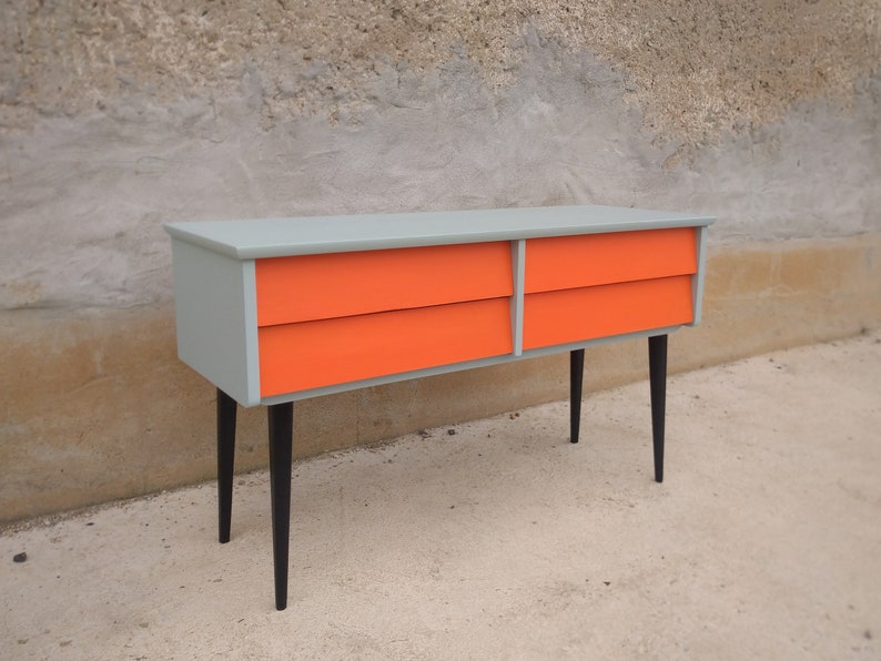 Das Bild zeigt eine original Vintage Kommode mit vier orange gestrichenen Schubladen. Der Korpus ist grün, und die langen Holzbeine sind schwarz gestrichen. Die Kommode wird leicht seitlich von links präsentiert. Blickfang mit Retro-Charme