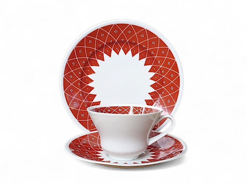 K&ouml;nnte beinhalten: Ein wei&szlig;es Tee-Set mit Tasse, Untertasse und Teller mit rotem geometrischem Muster. Das Muster zeigt ein Diamantgitter mit kleinen wei&szlig;en Punkten. Die Tasse und Untertasse haben das gleiche Design.