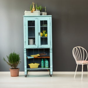 May include: A light blue vintage cabinet with wire mesh doors and shelves. The cabinet is filled with various items, including a yellow ceramic pot, a yellow ceramic bowl, and a few glass bottles. The cabinet is on wheels and has a wooden top.