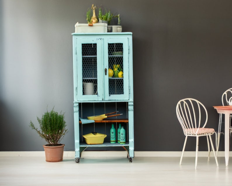 May include: A light blue vintage cabinet with wire mesh doors and shelves. The cabinet is filled with various items, including a yellow ceramic pot, a yellow ceramic bowl, and a few glass bottles. The cabinet is on wheels and has a wooden top.