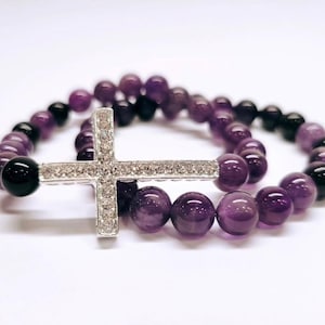 Könnte beinhalten: Zwei Armbänder mit Perlen und einem silbernen Kreuzanhänger. Die Armbänder haben lila und schwarze Perlen. Das Kreuz ist mit kleinen, klaren Kristallen besetzt. Die Armbänder werden auf einer weißen Oberfläche präsentiert.