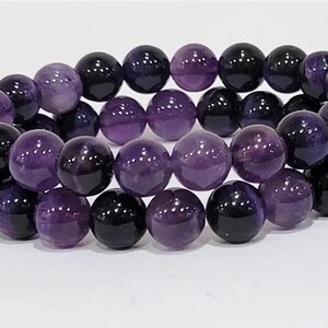 Könnte beinhalten: Ein Armband aus polierten, runden Amethystperlen. Die Perlen variieren in der Farbe von tiefem Lila bis fast Schwarz, wodurch ein Farbverlauf entsteht. Das Armband wird auf einer weißen Oberfläche präsentiert.