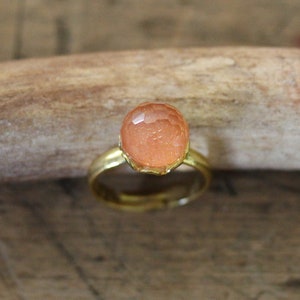 Könnte beinhalten: Ein goldfarbener Ring mit einem facettierten, pfirsichfarbenen Edelstein. Der Edelstein ist rund und in einer dekorativen, durchbrochenen Fassung gefasst. Der Ring ist schlicht und elegant.