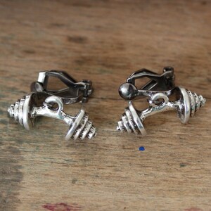 Puede incluir: Un par de pendientes de clip plateados con forma de mancuernas. Los pendientes tienen un diseño detallado y son perfectos para agregar un toque de estilo deportivo a cualquier atuendo.