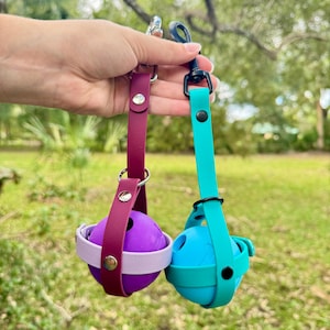 May include: Two dog treat dispensing balls with straps and clips. The ball on the left is purple and the ball on the right is blue.