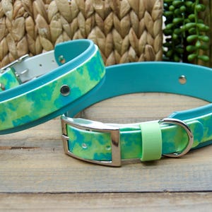 May include: Two waterproof dog collars featuring a vibrant tie-dye pattern in shades of turquoise and lime green. Each collar is constructed from a smooth, flexible material with a silver-toned metal buckle, a matching D-ring, and a light green keeper.