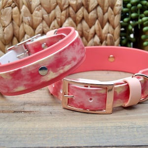 May include: Two pet collars featuring a pink and white tie-dye pattern. The collars are constructed from a smooth, flexible material with rose gold-toned metal hardware, including a buckle, a D-ring, and a central silver-toned metal rivet.