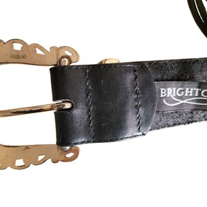 May include: A black leather belt with a decorative gold-tone buckle. The buckle has a scalloped edge design. The belt features the brand name "BRIGHTON" on a black tag.