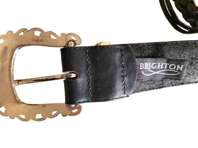 May include: A black leather belt with a decorative gold-tone buckle. The buckle has a scalloped edge design. The belt features the brand name "BRIGHTON" on a black tag.