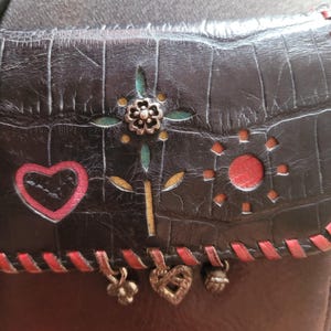 Puede incluir: Bolso bandolera de cuero negro con textura de cocodrilo en relieve. La solapa presenta un dise&ntilde;o floral, un coraz&oacute;n rojo y un sol. Costuras rojas y abalorios met&aacute;licos a&ntilde;aden detalles. El bolso tiene una correa para el hombro.