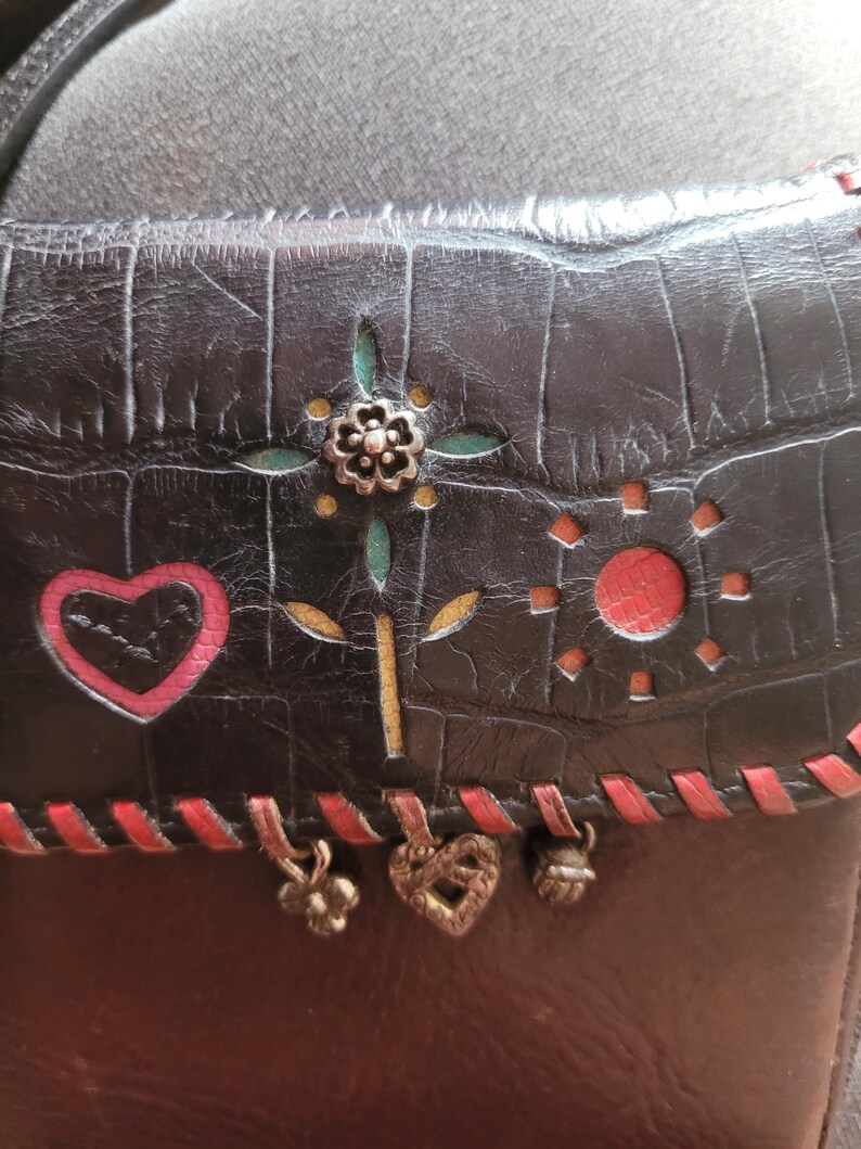 Puede incluir: Bolso bandolera de cuero negro con textura de cocodrilo en relieve. La solapa presenta un dise&ntilde;o floral, un coraz&oacute;n rojo y un sol. Costuras rojas y abalorios met&aacute;licos a&ntilde;aden detalles. El bolso tiene una correa para el hombro.