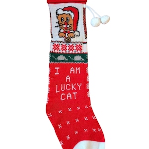 May include: A red and white Christmas stocking with a knitted design. The stocking features a cat wearing a Santa hat, the text "I AM A LUCKY CAT", and a white toe. A small bell hangs below the cat.