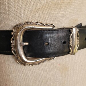 May include: Close-up of a black leather belt with a silver-colored, ornate buckle. The buckle has a decorative, scrolled design. The belt is a classic accessory, suitable for various outfits.