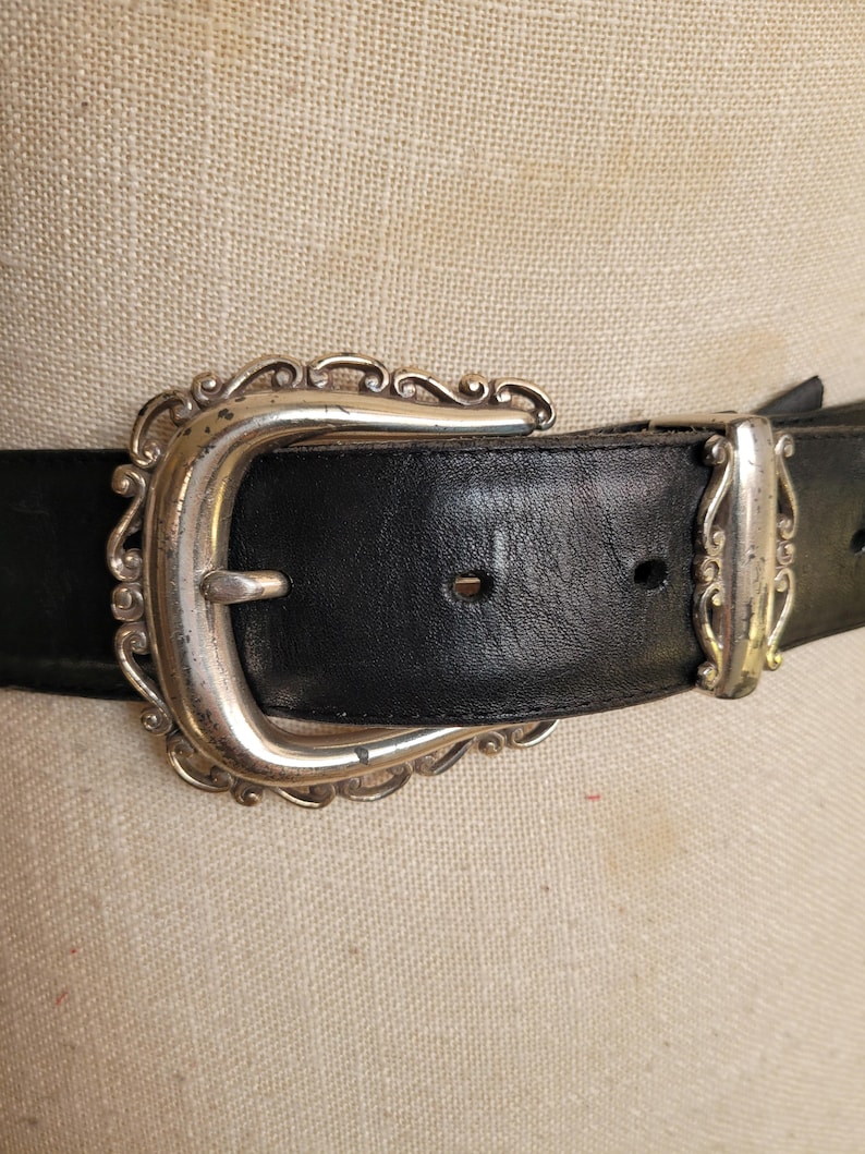 May include: Close-up of a black leather belt with a silver-colored, ornate buckle. The buckle has a decorative, scrolled design. The belt is a classic accessory, suitable for various outfits.
