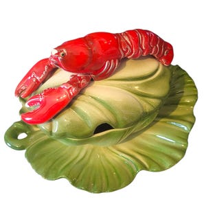 May include: A ceramic soup tureen with a bright red lobster lid. The tureen is shaped like a green cabbage leaf with a matching base. The lobster has detailed claws and a textured body. The tureen has a small handle and a spoon opening.