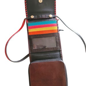 Puede incluir: Bolso bandolera de cuero marr&oacute;n abierto, con interior negro y ribete cosido en rojo. El bolso tiene un compartimento inferior con cremallera, una ventana de identificaci&oacute;n transparente y ranuras para tarjetas de colores. La correa es negra y roja.