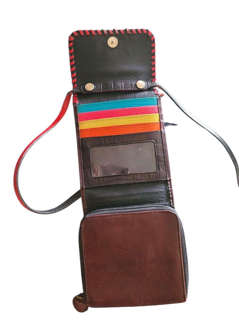 Puede incluir: Bolso bandolera de cuero marr&oacute;n abierto, con interior negro y ribete cosido en rojo. El bolso tiene un compartimento inferior con cremallera, una ventana de identificaci&oacute;n transparente y ranuras para tarjetas de colores. La correa es negra y roja.