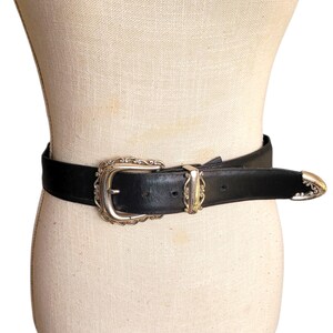 May include: A black leather belt with a silver-toned buckle and matching end caps. The buckle has an ornate, vintage design. The belt is displayed on a mannequin, showcasing its style and detailing.