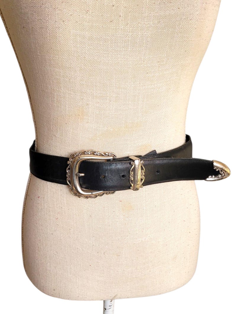 May include: A black leather belt with a silver-toned buckle and matching end caps. The buckle has an ornate, vintage design. The belt is displayed on a mannequin, showcasing its style and detailing.