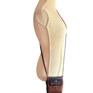 Puede incluir: Un peque&ntilde;o bolso bandolera marr&oacute;n y azul con una larga correa negra. El bolso presenta una solapa decorativa con flores y un coraz&oacute;n bordados. La parte inferior del bolso es marr&oacute;n y la superior es azul.