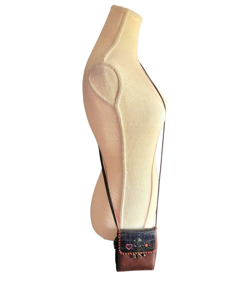 Puede incluir: Un peque&ntilde;o bolso bandolera marr&oacute;n y azul con una larga correa negra. El bolso presenta una solapa decorativa con flores y un coraz&oacute;n bordados. La parte inferior del bolso es marr&oacute;n y la superior es azul.