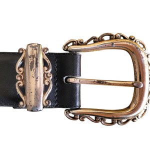 May include: Close-up of a black leather belt with a decorative silver buckle. The buckle has an ornate design with a silver center and a gold-colored frame. The belt is a classic accessory.