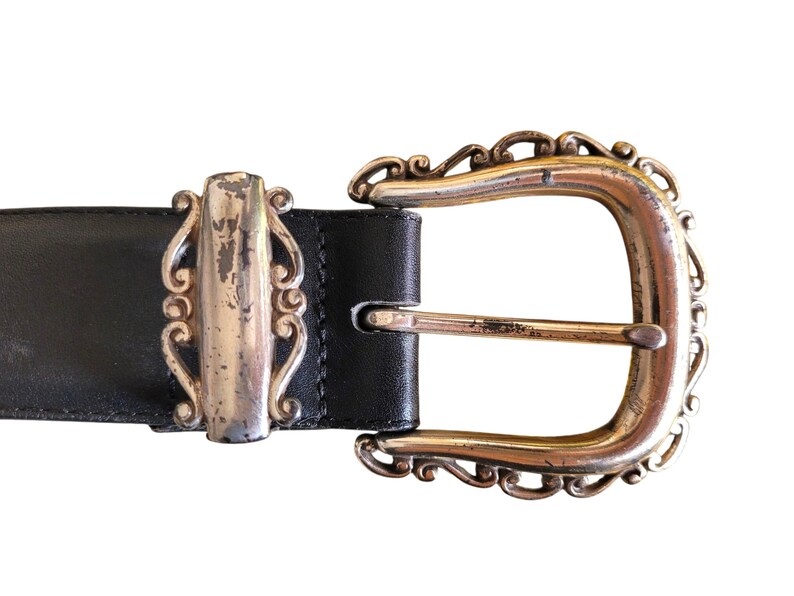 May include: Close-up of a black leather belt with a decorative silver buckle. The buckle has an ornate design with a silver center and a gold-colored frame. The belt is a classic accessory.