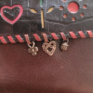 Puede incluir: Una cartera de cuero de dos tonos con una secci&oacute;n superior negra y una inferior marr&oacute;n. La secci&oacute;n negra presenta dise&ntilde;os recortados de un coraz&oacute;n, un sol y una flor. Cuelgan dijes decorativos de metal del borde inferior.