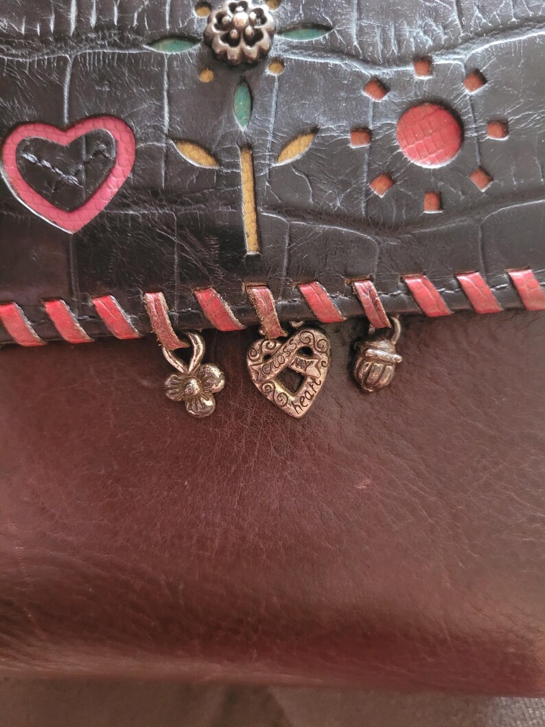 Puede incluir: Una cartera de cuero de dos tonos con una secci&oacute;n superior negra y una inferior marr&oacute;n. La secci&oacute;n negra presenta dise&ntilde;os recortados de un coraz&oacute;n, un sol y una flor. Cuelgan dijes decorativos de metal del borde inferior.