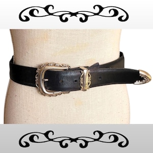May include: Black leather belt with a silver-toned buckle and tip. The buckle has an ornate, scrolled design. The belt is displayed on a mannequin. A decorative black and white design is above and below the belt.