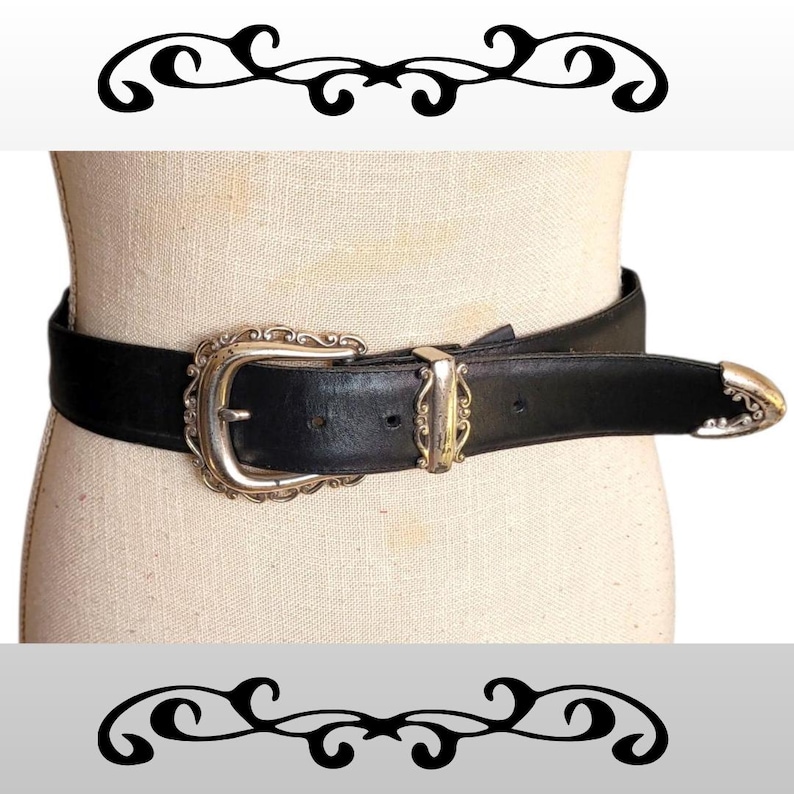 May include: Black leather belt with a silver-toned buckle and tip. The buckle has an ornate, scrolled design. The belt is displayed on a mannequin. A decorative black and white design is above and below the belt.