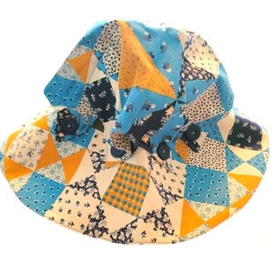 May include: A patchwork sun hat with a wide brim and a rounded crown. The hat features a variety of fabric patterns in shades of blue, yellow, and white, with dark blue buttons. The hat is designed for sun protection.