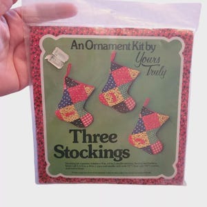 May include: A kit for making three Christmas stockings. The package features a green label with the text "An Ornament Kit by Yours Truly" and "Three Stockings." The stockings are patchwork with red, blue, and yellow fabrics. The package is in a clear plastic bag.
