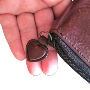 Puede incluir: Primer plano de una cartera de cuero marr&oacute;n con un tirador de cremallera en forma de coraz&oacute;n. La cartera tiene una cremallera negra y una superficie texturizada. El tirador en forma de coraz&oacute;n est&aacute; unido a un anillo de metal.