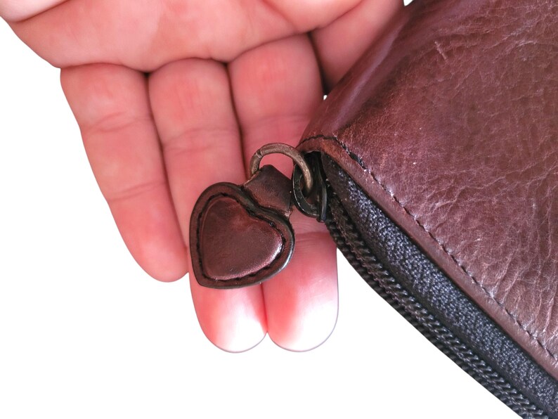 Puede incluir: Primer plano de una cartera de cuero marr&oacute;n con un tirador de cremallera en forma de coraz&oacute;n. La cartera tiene una cremallera negra y una superficie texturizada. El tirador en forma de coraz&oacute;n est&aacute; unido a un anillo de metal.