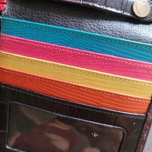 Puede incluir: Una cartera de cuero negro y marr&oacute;n con un interior colorido. La cartera presenta ranuras para tarjetas en turquesa, rosa, amarillo y naranja. Tiene una ventana de identificaci&oacute;n transparente y dos cierres a presi&oacute;n en forma de coraz&oacute;n.