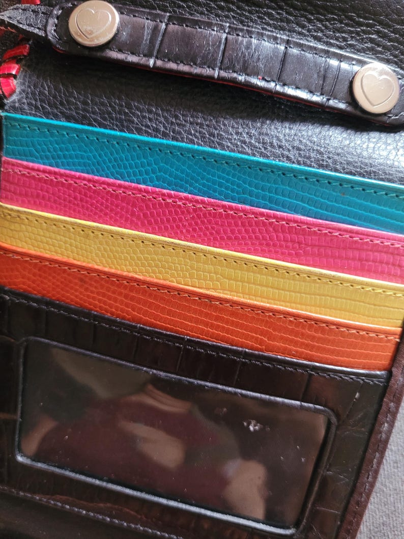 Puede incluir: Una cartera de cuero negro y marr&oacute;n con un interior colorido. La cartera presenta ranuras para tarjetas en turquesa, rosa, amarillo y naranja. Tiene una ventana de identificaci&oacute;n transparente y dos cierres a presi&oacute;n en forma de coraz&oacute;n.