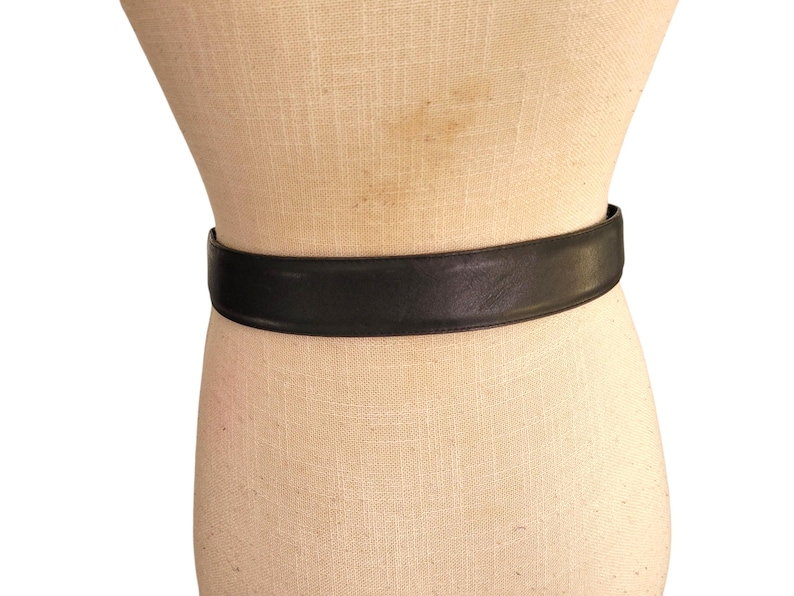 May include: A black leather belt is wrapped around a mannequin. The belt is a solid, dark color and appears to be made of smooth leather. The belt is a simple, classic design.