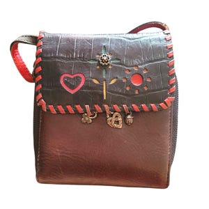 Puede incluir: Un peque&ntilde;o bolso bandolera de cuero marr&oacute;n con una solapa gris oscuro texturizada. La solapa presenta costuras rojas, dise&ntilde;os recortados y un adorno floral. El bolso tiene una correa roja y abalorios decorativos.