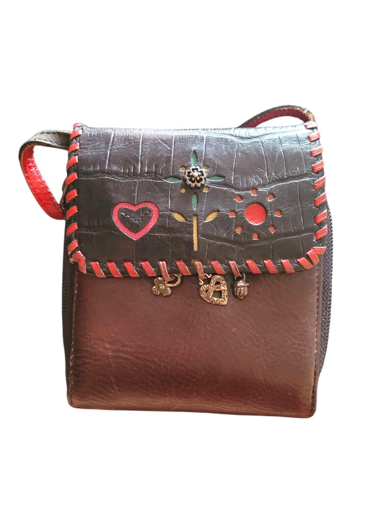 Puede incluir: Un peque&ntilde;o bolso bandolera de cuero marr&oacute;n con una solapa gris oscuro texturizada. La solapa presenta costuras rojas, dise&ntilde;os recortados y un adorno floral. El bolso tiene una correa roja y abalorios decorativos.