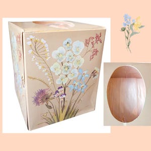 May include: A beige tissue box featuring a floral design with light blue, white, purple, and pink flowers. The box has an oval opening on top for tissue dispensing. A watercolor illustration of flowers is also present.