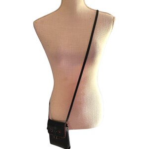 Puede incluir: Un bolso bandolera negro con una correa larga y fina cuelga de un maniqu&iacute; beige. El bolso tiene un cierre de solapa con costuras decorativas rojas y negras. El maniqu&iacute; es de color neutro.