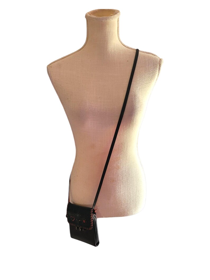 Puede incluir: Un bolso bandolera negro con una correa larga y fina cuelga de un maniqu&iacute; beige. El bolso tiene un cierre de solapa con costuras decorativas rojas y negras. El maniqu&iacute; es de color neutro.