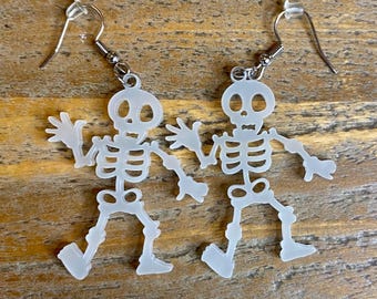 May include: A pair of skeleton earrings made of white acrylic. The earrings are shaped like skeletons with their arms raised in a waving motion. The earrings are attached to silver hooks.