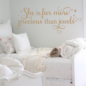 May include: White bed with white bedding and a white throw blanket. The wall behind the bed has a gold vinyl decal that reads "She is far more precious than jewels".