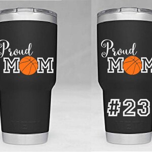 May include: Two black stainless steel tumblers with a white design that says "Proud Mom" with a basketball graphic. One tumbler has the number "23" on the bottom.