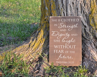 May include: A brown wooden sign with white text that reads "She is clothed in Strength and Dignity and she laughs without fear of the future. Proverbs 31:25". The sign is leaning against a tree trunk.