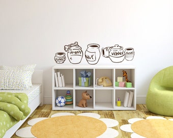 May include: A set of Winnie the Pooh honey pot wall decals in brown. The decals feature a variety of honey pots with the word "Hunny" written on them. The decals are displayed on a white wall above a white bookshelf with a green armchair and a bed with green and white bedding.
