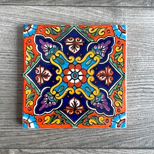 May include: A square ceramic tile with a colorful, intricate design. The tile features a central motif of a flower with four petals, surrounded by swirling patterns and smaller floral elements. The colors used are vibrant and include shades of blue, red, yellow, green, and purple.